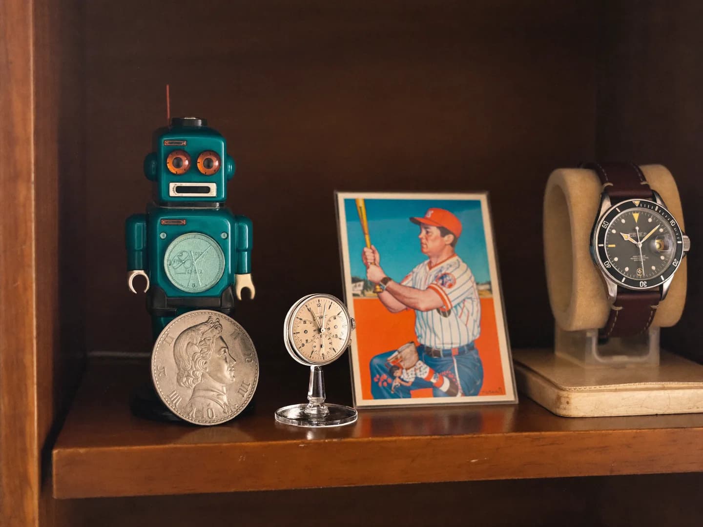 A silver coin, vintage tin robot, baseball card, and diver watch arranged on a wooden shelf
